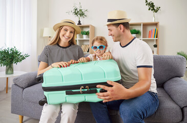 Portrait of happy cheerful young parents with child daughter sitting on sofa with packed suitcase...