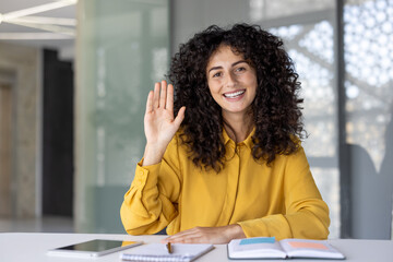 Woman sitting inside office at workplace looking at camera, waving hello and talking to colleagues...