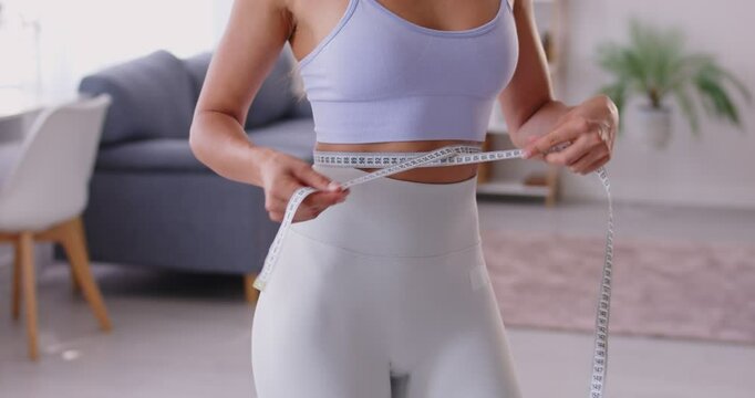 Cropped portrait of faceless slim sporty woman wearing sportswear measure the waist with measuring tape standing at home. Slimming, sport, diet and weight control concept. 4k video.