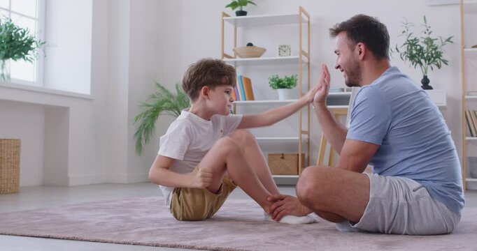 Father helps his child son do fitness exercises during a family sports workout at home. Happy dad holds the little boy's feet and gives him a high five every time he sits up