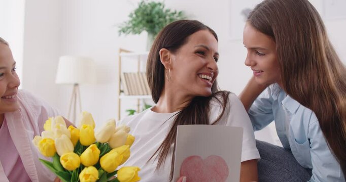 Smiling loving teen daughter sisters greeting happy mother with anniversary presenting bouquet of flowers, card, happy mom and grownup children in hug embraces to celebrate birthday at home together