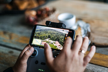 Close-up of hands using a smartphone and drone controller to navigate and view live aerial footage of a scenic landscape with forest and houses. © irissca