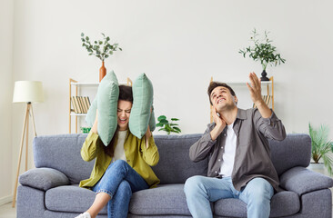 Couple sits on the sofa at home living room, covering ears due to loud noise from neighbors. They...