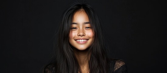 Fototapeta premium Asian girl smiling gently against a black studio background with soft lighting and ample copyspace for text, featuring long dark hair and golden highlights.