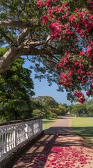 Fototapeta premium Pathway with scattered pink petals under a tree in full bloom, surrounded by lush greenery and a white railing on a sunny day