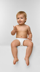 A baby girl, toddler in a diaper curiously sitting and gesturing on a white cube, looking with...