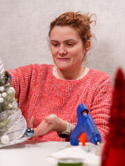 A woman in a cozy sweater meticulously crafts holiday decorations using a glue gun. Various craft...