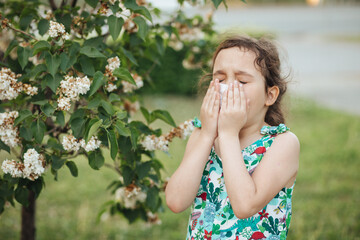 Little kid girl with seasonal allergy gets cold and blows her nose outdoor nearby blooming trees. Healthcare, pollen and flowering allergy in spring concept. World Allergy Day.