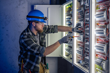 A male electrician works in a switchboard in overalls against the backdrop of emergency lighting.