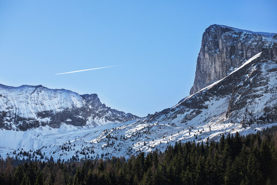 Montagne enneig&eacute;e dominant une vall&eacute;e bois&eacute;e et enneig&eacute;e avec avion passant dans le ciel bleu d'hiver.