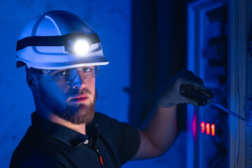 A male electrician works in a switchboard in overalls against the backdrop of emergency lighting.