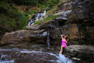 A beautiful girl in a pink dress sits on a stone near Ravana waterfall, Sri Lanka