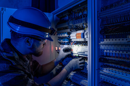 A male electrician works in a switchboard in overalls against the backdrop of emergency lighting.