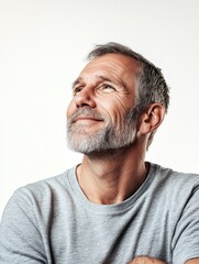 Obraz premium Mature man with gray beard looking upwards with a joyful expression, light gray shirt against soft white background, perfect for personalized text overlay.
