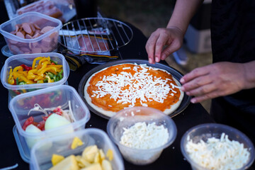 Chef hands is putting sliced cheese on pizza with ingredients in boxes on a black table.