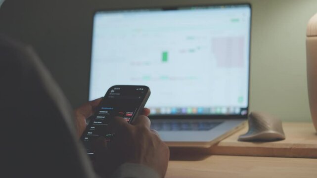 A person is using a laptop and a cell phone to check the stock market. The laptop screen shows a graph of the stock market, while the cell phone is being used to access additional information