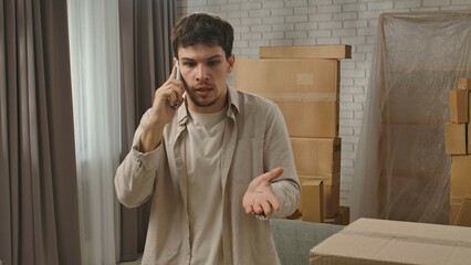 Young couple moving into new apartment, space filled with cardboard boxes, man stands in the room of new flat and talks on smartphone, frustrated negative expression.