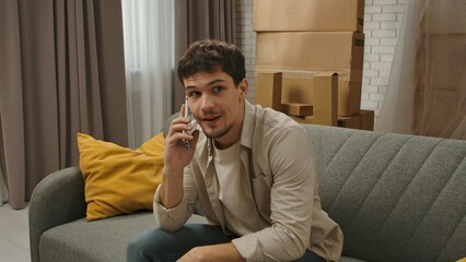 Young couple moving into new apartment, space filled with cardboard boxes, man sits on the sofa in room of new apartment talks on smartphone, happy positive expression.