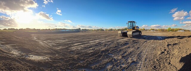 Construction machinery prepares land for development in an open field under a bright sky during sunset