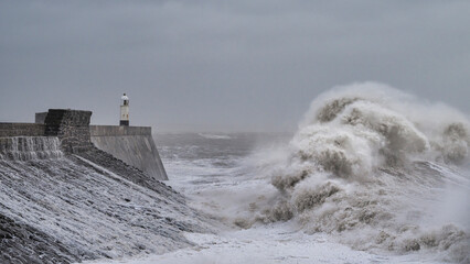 A huge winter storm hitting the coast of the UK. Giant waves crash into a breakwater, with a lighthouse. The sky is grey and moody