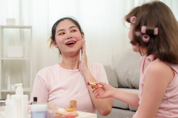 Asian mother admiring playful makeup attempt by daughter during joyful beauty activity at home. Heartwarming interaction representing love, trust, and family bonding in a cheerful setting.