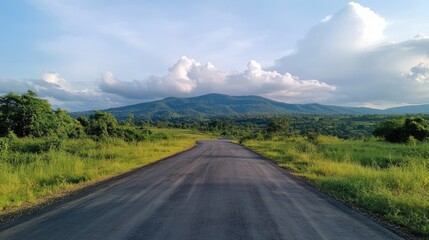 Naklejka premium Asphalt road winding through lush greenery under a scenic sky with mountains and clouds in the background