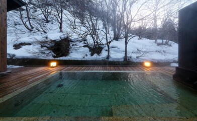 A calm, relaxing winter scene of a chartered open-air hot bath of natural thermal spring, which is lighted up at dusk and located in the forest on a snowy hillside, in an Onsen Ryokan in Iwate, Japan