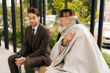 Kind businessman in suit listening with empathy to homeless elderly man talking sitting on urban bench. Compassionate entrepreneur with coffee engaging in conversation with homeless senior citizen