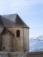 A historic church in Briançon, France, with aged stone walls and a bell tower topped with a cross. Snowy mountains in the background enhance the scenic charm.