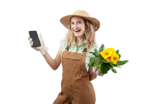 A smiling woman wearing gardening clothes, holding a pot with blooming yellow primrose flowers, isolated on a white background, shows a smartphone. Online flower shopping for spring and garden work