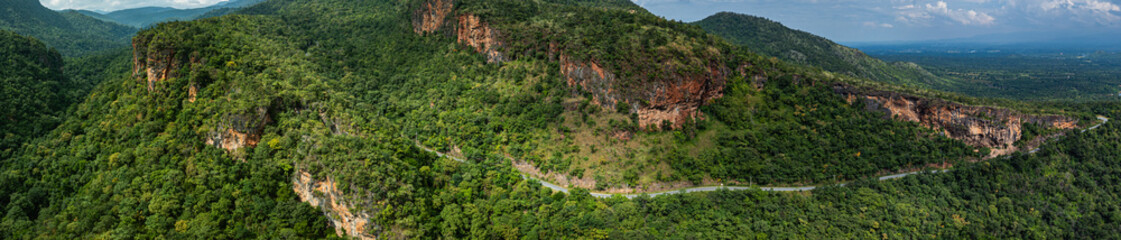 Aerial panoramic view of lush green hills and valleys intersected by a winding mountain road. The landscape features rugged cliffs, dense forests, and a distant horizon under a partly cloudy sky.