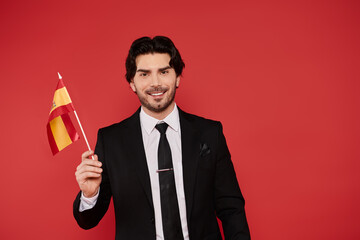 Young man in a suit smiles while holding a Spanish flag against a bold red background