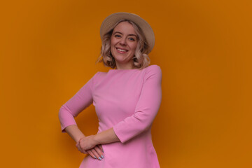 Smiling woman in a pink dress and straw hat posing confidently against a vibrant orange background, exuding joy, style, and charm in a modern lifestyle portrait.