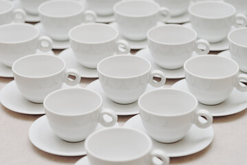 white empty porcelain cups on saucers with spoons in several rows on white tablecloth, serving tables for banquet in restaurant or cafe, pattern, background