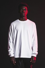 Obraz premium A young man stands confidently in a white long sleeve T-shirt mockup against a dark background with red lighting. His expression is serious, and the contrast highlights his features.