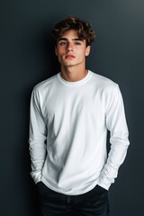 Obraz premium A young man with curly hair wearing a white long sleeve T-shirt mockup, standing against a dark wall. He has a relaxed expression and hands in his pockets.