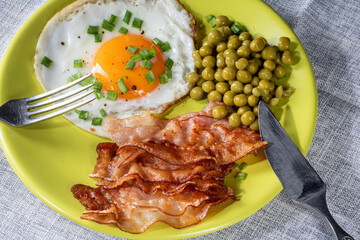 Fried egg with bacon and green peas on a plate, top view