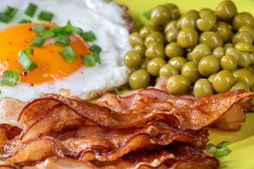 Fried eggs with bacon and green peas on a plate, front view