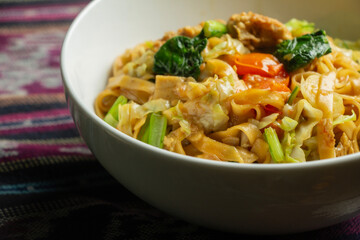 Thai Drunken Noodles with egg and sausage combined in a white bowl, negative space on a traditional cloth base