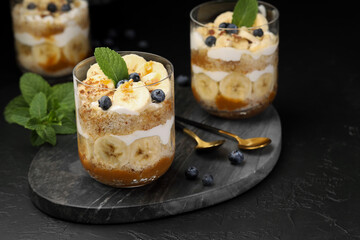 Tasty trifle dessert. Sponge cake, blueberries, banana and whipped cream in glasses on black table