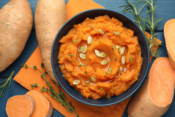 Tasty mashed sweet potato with pumpkin seeds in bowl, fresh vegetables, rosemary and thyme on blue wooden table, flat lay