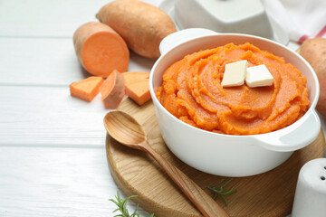 Tasty mashed sweet potato with butter in bowl, spoon and fresh vegetables on white wooden table, closeup