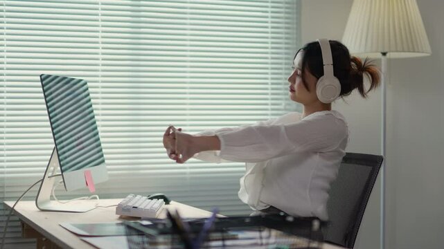Office professional wearing headphones stretching arms while sitting at workspace, pausing from work with relaxed, ergonomic movement near computer and window