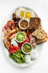 A delicious breakfast spread featuring sunny-side-up eggs, crispy bacon, sausages, assorted vegetables, toast, and dips like guacamole and tuna. Perfect for a hearty meal.