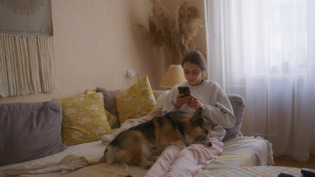 young woman in a cozy home setting lies on the couch with her Welsh Corgi dog. She scrolls through social media on her smartphone while the dog sleeps peacefully on her legs