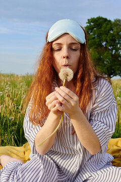 Relaxed woman wearing a sleep mask blowing on a dandelion in nature