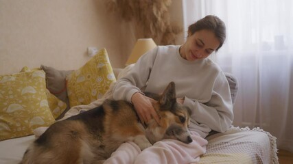 woman relaxes on couch with Corgi lying adorably on her legs. She gently pets dog, hugging and talking to it with love. heartwarming scene captures bond between pet and owner in serene home setting.