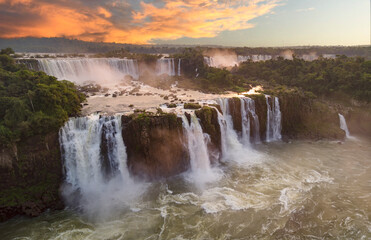 Fototapeta premium Majestic Iguazu Falls with powerful cascades flowing amidst lush green vegetation, illuminated by a vibrant sunset sky