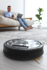 Young man with cat using laptop at home, focus on robotic vacuum cleaner