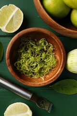 Lime zest, fresh fruits and zester tool on green wooden table, flat lay